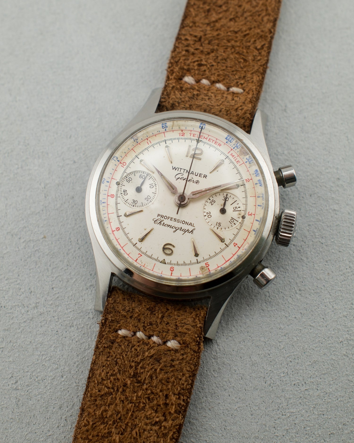 Vintage steel watch with a chronograph stopwatch dial and blue and red details on it surface. It had a brown suede strap.