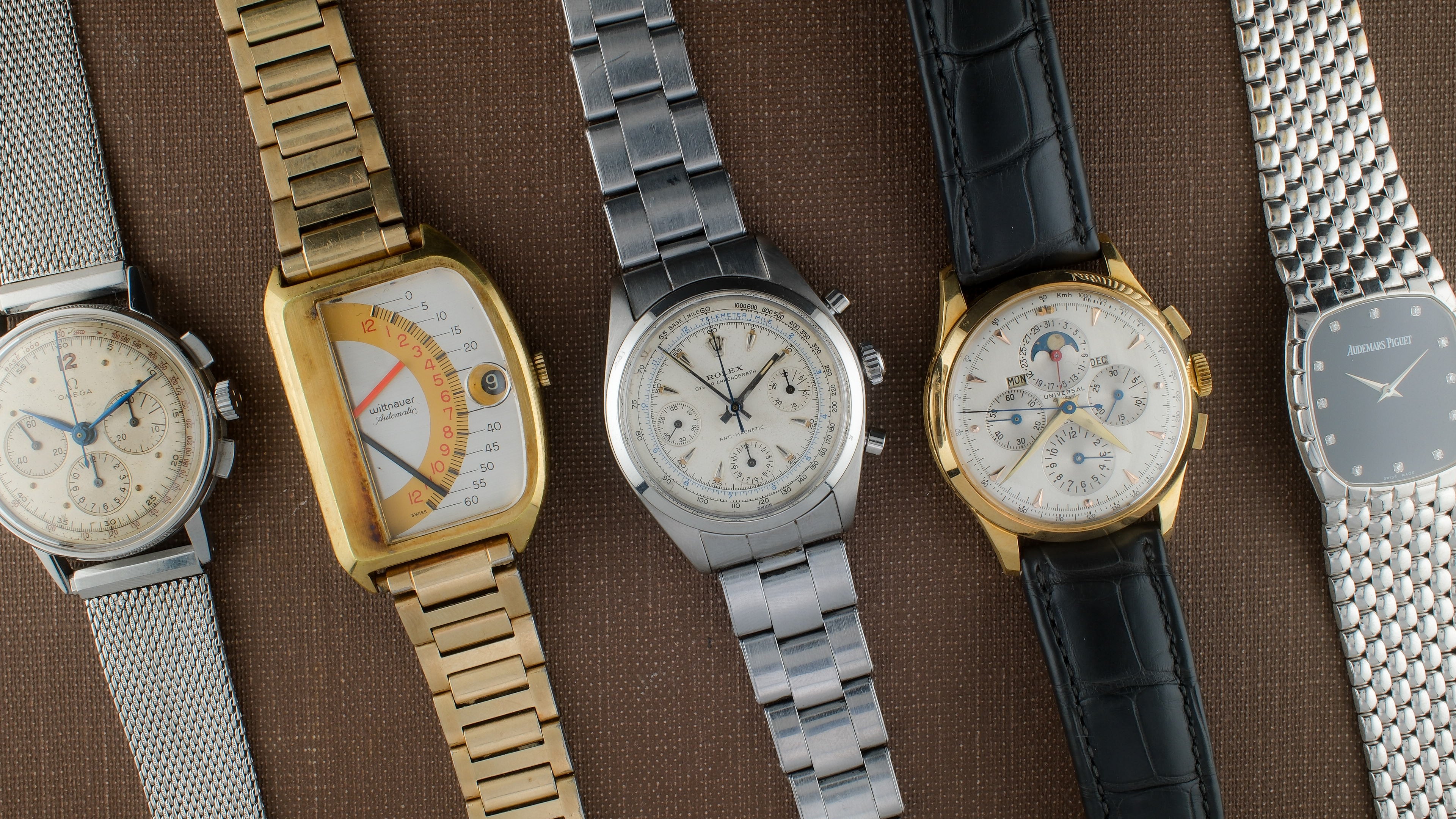 A selection of rare vintage watches. A steel omega chronograph, a gold rectangular watch, a white and blue dial rolex chronograph, a gold universal genève with three smaller dials and two small rectangular registers that display different information, and a white gold audemars piguet with a bracelet that looks like snake skin.