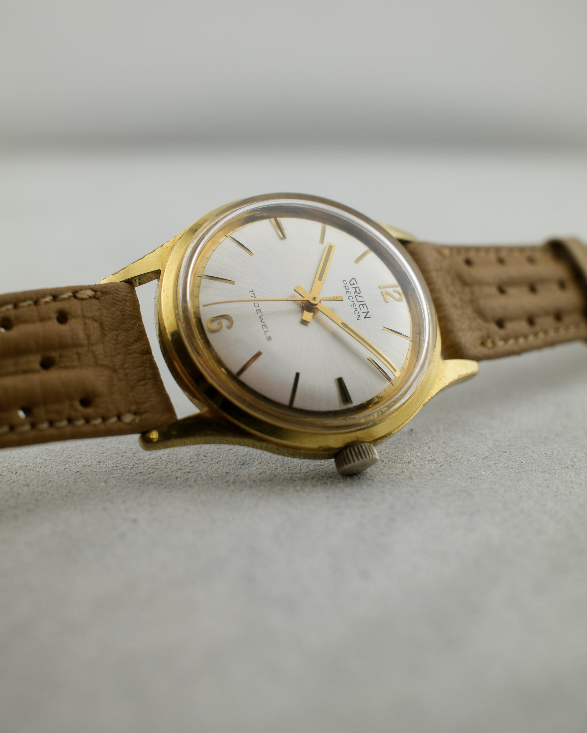 1960's Gruen gold plated watch with white dial, gold hands, and light brown strap sitting on its side.