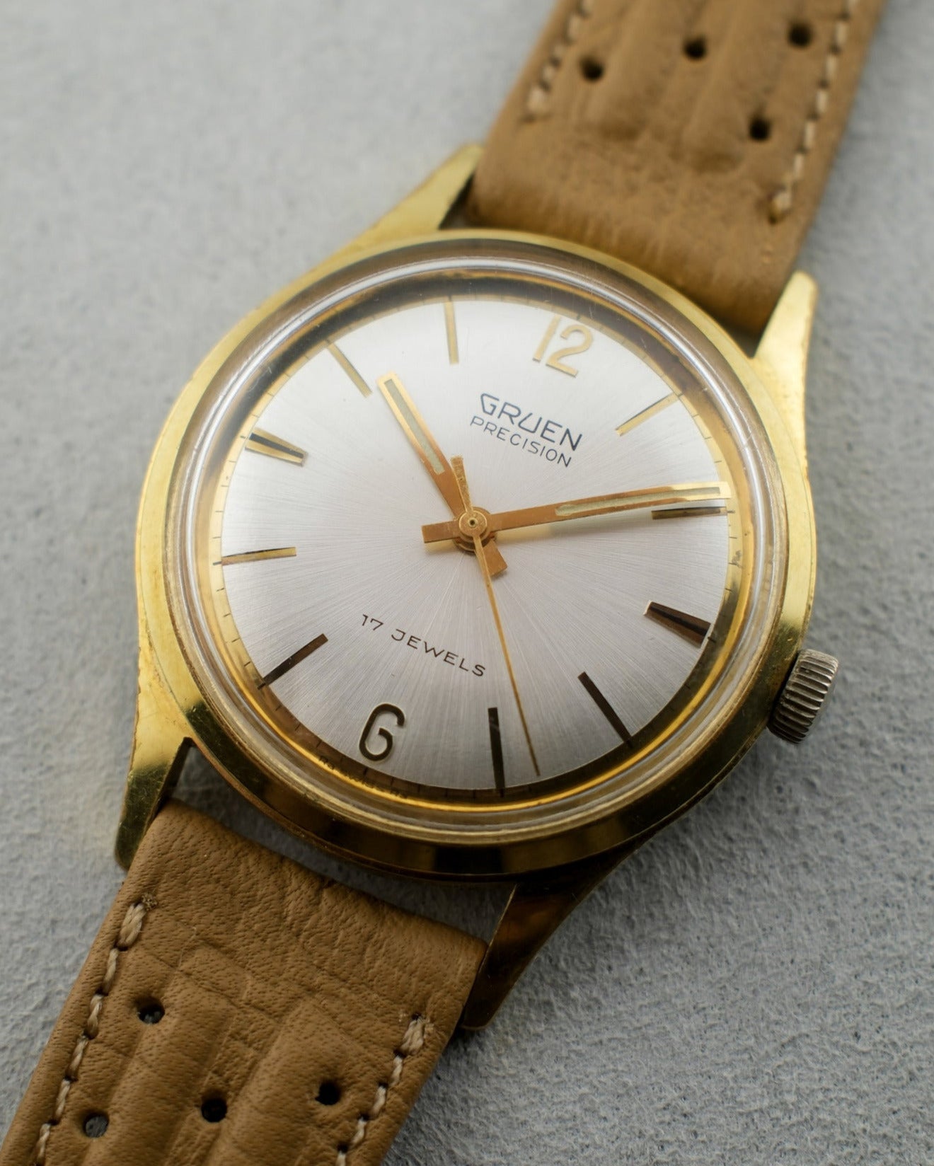 1960's Gruen gold plated watch with white dial, gold hands, and light brown strap.