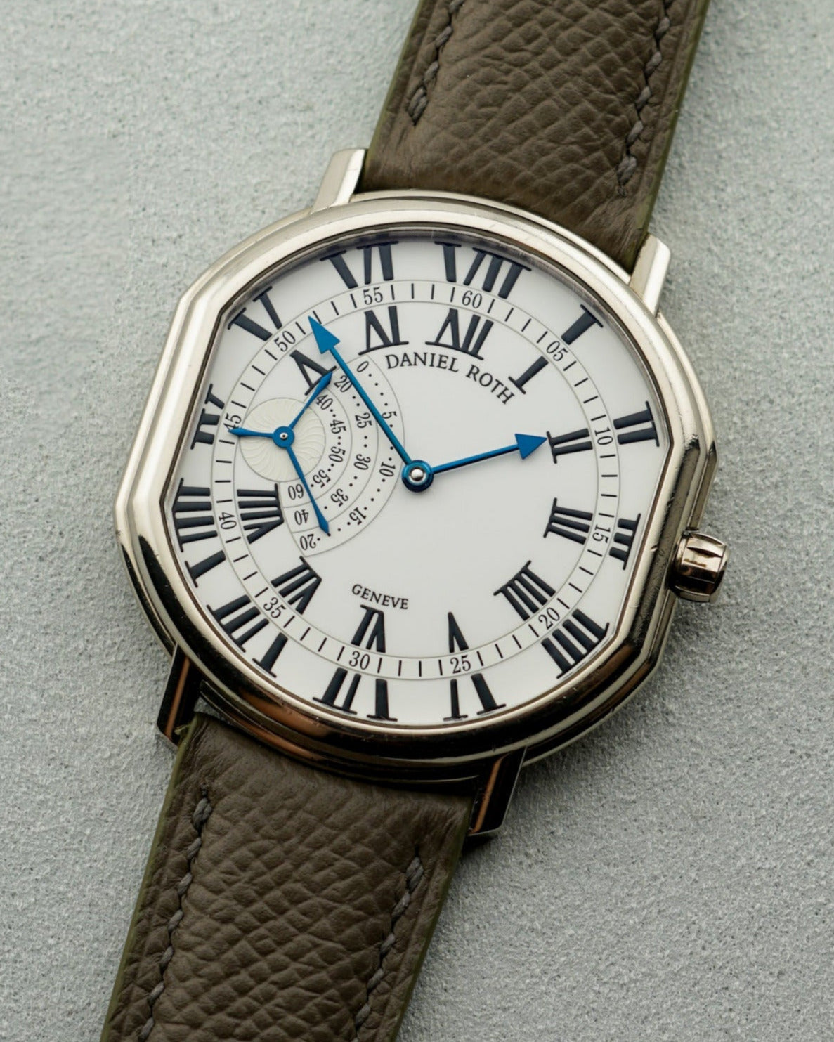 Daniel Roth watch with bold white dial, roman numeral hour markers, blue arrow hands, and a small seconds complication at 9 o'clock. It wears a dark grey, almost brown colored leather strap.
