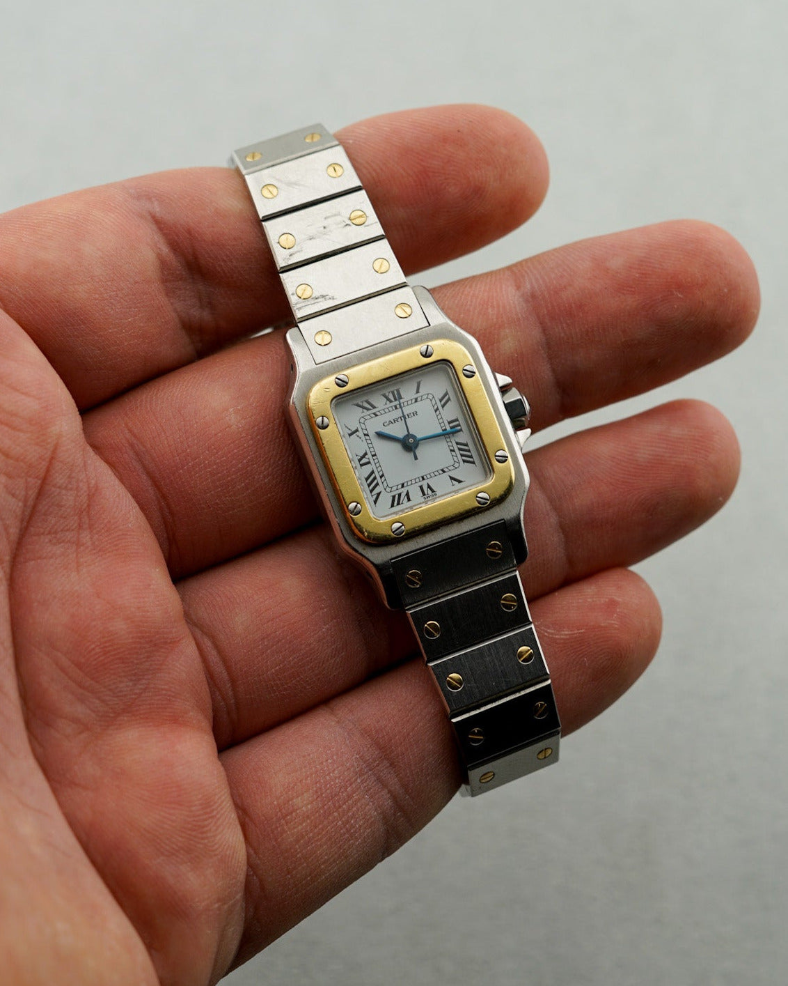 Small square watch with gold and silver accents held in a hand against a plain background