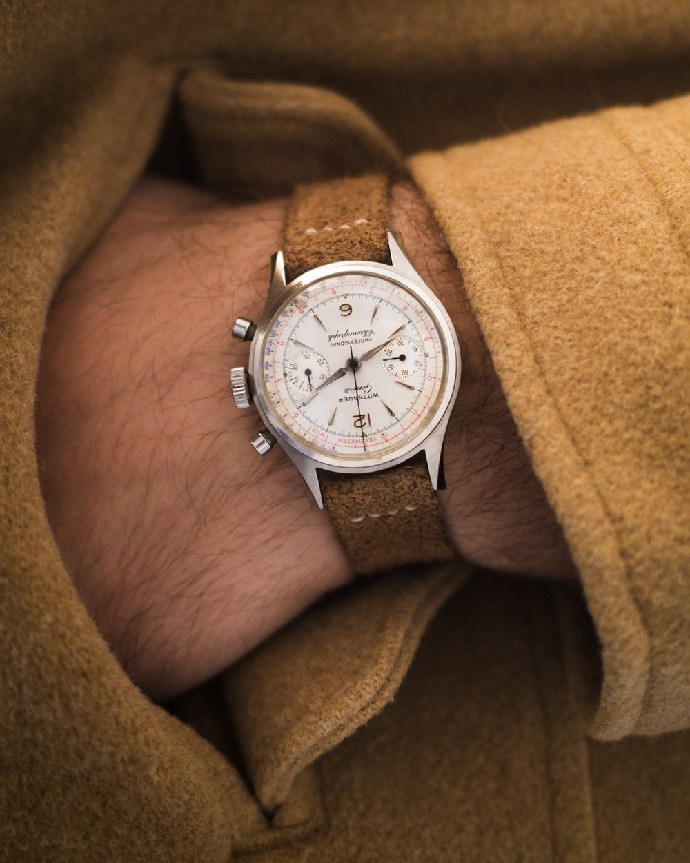 Vintage Wittnauer chronograph wristwatch with brown leather strap worn on a wrist, set against a brown coat background.